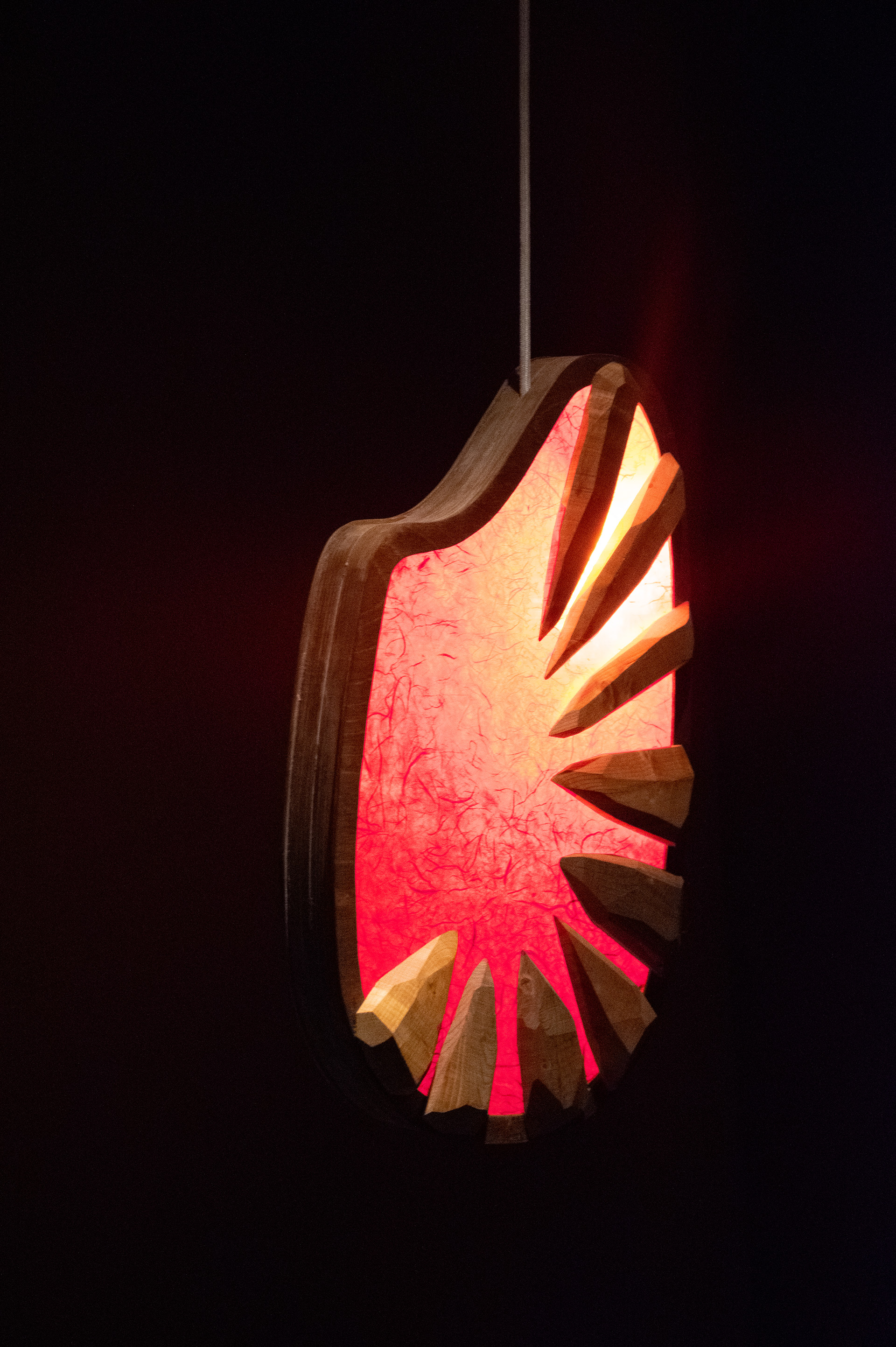 A rounded red paper and wood lamp with jagged edges pointing inwards.. the lamp is displayed in darkness at a 3/4 view.