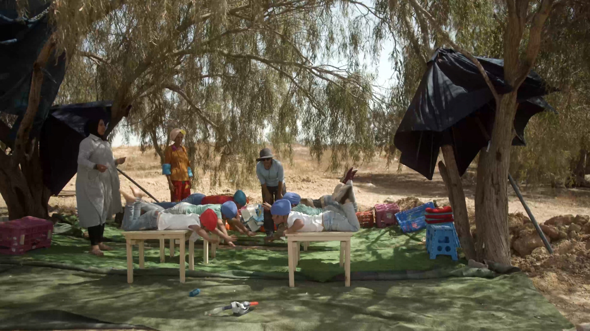 Image from video still, six children in blue swim caps laying on wooden tables practicing swimming in the desert under trees, surrounded by three women teaching them.